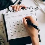 Top view of hands using a planner with coffee and accessories, focused on May 2021 calendar.