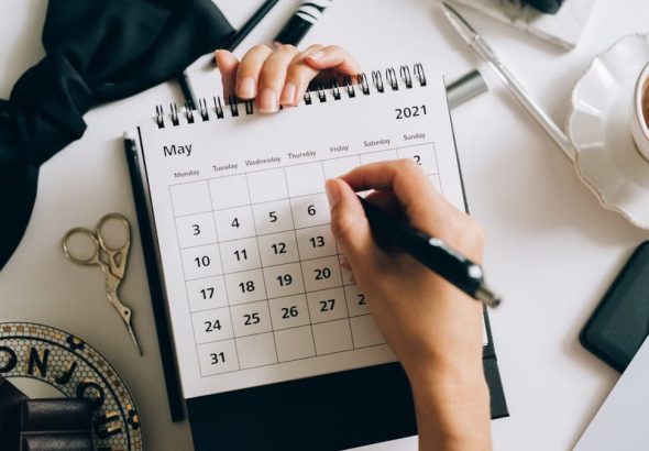 Top view of hands using a planner with coffee and accessories, focused on May 2021 calendar.